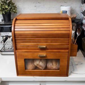May include: A two-tier wooden bread box with a roll-top upper door and a lower drawer featuring a horizontal rectangular window made of ribbed glass. The unit has a warm brown finish and is equipped with two rectangular metallic handles.
