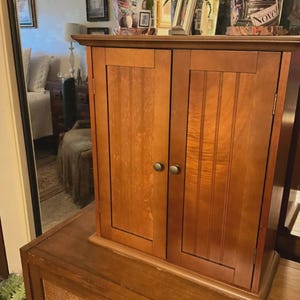 May include: A medium-brown wooden cabinet featuring two vertical doors with recessed panels and small, round, metallic knobs. The cabinet has a flat top surface and a rectangular frame, showcasing a smooth, polished wood grain finish throughout.
