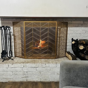 May include: A fireplace with a burning fire behind a gold-colored screen. The brick hearth is painted white. Black fireplace tools stand to the left. A log holder with firewood is on the right.