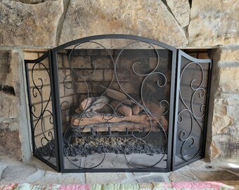 May include: Black metal fireplace screen with decorative scrollwork and mesh panels. The screen is tri-fold and protects the fireplace opening. The fireplace has a brick interior and a stone surround.