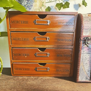 May include: A small, wooden storage cabinet with four drawers. Each drawer is labeled with the words "MERCERIE" and "BOUTON" in black text. The cabinet has a warm, brown finish and metal label holders.