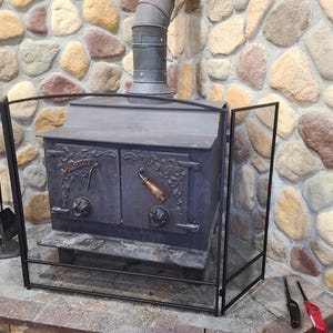 May include: A black metal wood-burning stove with a mesh screen in front. The stove has two doors with decorative handles and the word "Arster" on the front. A set of fireplace tools and a stone wall are in the background.