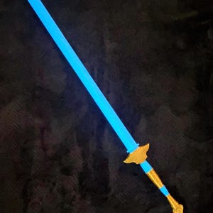 May include: A toy sword featuring a long, glowing light blue blade. The hilt consists of a gold-colored crossguard with a wing-like design, a light blue grip, and a gold-colored pommel at the base of the handle, all set against a dark background.