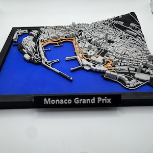 Peut inclure: Une maquette imprimée en 3D du circuit du Grand Prix de Monaco, avec une représentation détaillée de la ville et du tracé de la course. Le circuit est mis en évidence en orange sur un paysage urbain gris et noir, sur fond bleu dans un cadre noir. Le texte "Monaco Grand Prix" est affiché.
