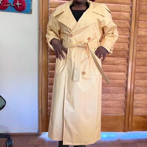 Könnte beinhalten: Ein hellgelber Trenchcoat mit passendem Gürtel und Knopfverschlüssen. Der Mantel hat ein klassisches doppelreihiges Design und ist knielang. Die Person trägt dunkelgrüne Stiefel. Der Hintergrund umfasst Holzläden und eine Wand mit einem dekorativen Element.