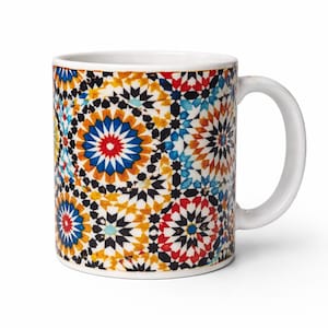 May include: White ceramic mug featuring a colorful geometric pattern in shades of blue, red, yellow, and black. The design is reminiscent of mosaic tiles. The mug has a white handle and a capacity of approximately 12 ounces.