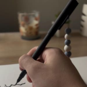 Puede incluir: Una mano sostiene un bolígrafo negro, creando una línea curva sobre papel blanco. La punta del bolígrafo toca el papel, dejando una marca oscura. El fondo está borroso, con una bebida y cuentas decorativas visibles.