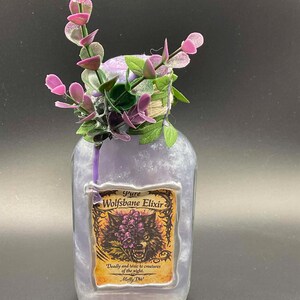 May include: A clear glass bottle filled with a swirling light purple liquid, topped with artificial purple and green foliage. The label reads Pure Wolfsbane Elixir, Deadly and toxic to creatures of the night, with an illustration of a wolf head.