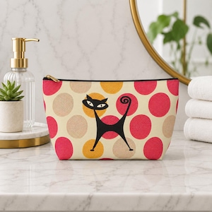 May include: A beige cosmetic bag featuring a black cat illustration and a pattern of pink, yellow, and beige polka dots. The bag has a black zip and trim. The bag is on a marble surface.