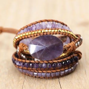 Puede incluir: Una pulsera envolvente multicapa con una gran piedra de amatista ovalada facetada. La pulsera incorpora pequeñas cuentas moradas, detalles dorados y correas de cuero marrón. El diseño es bohemio y artesanal.