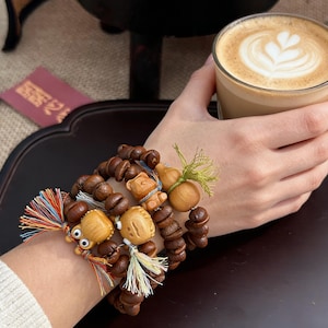 Könnte beinhalten: Eine Sammlung von Holzperlenarmbändern mit verschiedenen Anhängern, darunter eine Eule, ein Bär und eine Kalebasse. Die Armbänder sind braun mit bunten Quastenakzenten. Eine Hand hält ein Glas Kaffee mit Latte Art.