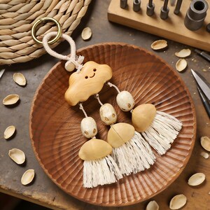 May include: A wooden cloud-shaped keychain with a smiling face, cream-colored tassels, and a gold-colored ring. The keychain is displayed on a carved wooden bowl with pistachio shells scattered around it.