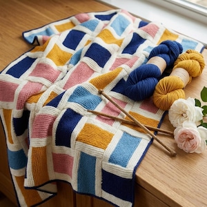 Puede incluir: Una manta tejida a mano, colorida, con un patrón geométrico en tonos azules, rosas, amarillos y blancos. Dos madejas de hilo, una azul y otra amarilla, y dos agujas de tejer de madera están junto a la manta. También hay algunas rosas.