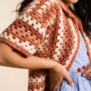 May include: A crocheted, short-sleeved cardigan in a granny square pattern. The color palette includes shades of brown, cream, and orange. The garment has a collar and is worn over a light blue dress with floral embroidery.