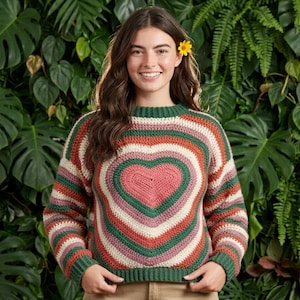 May include: A long-sleeved crochet sweater featuring a large central pink heart motif surrounded by concentric stripes in shades of dark green, cream, and rust orange. The garment has a ribbed crew neckline and ribbed cuffs in a solid dark green yarn.