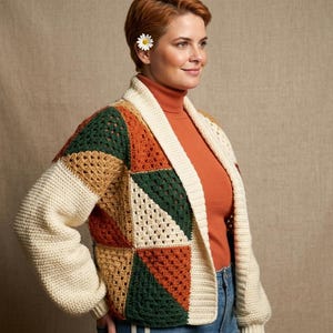 May include: A patchwork crochet cardigan in cream, green, orange, and tan. The open-front sweater has long sleeves and a shawl collar. The model is wearing a rust-coloured turtleneck and blue jeans. A daisy is tucked behind the ear.