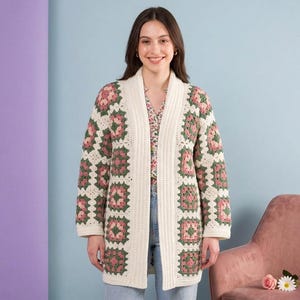 Könnte beinhalten: Ein cremefarbener, langer Häkelcardigan mit einem Blumenmuster in Rosa und Grün. Der Cardigan wird offen getragen und zeigt ein Oberteil mit Blumenmuster und blaue Jeans. Der Hintergrund ist eine Mischung aus Lila und Blau.