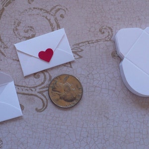 May include: Three white envelopes of different sizes, one with a red heart on the front. The envelopes are on a light brown surface with a pattern.