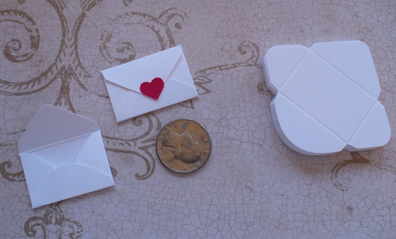 May include: Three white envelopes of different sizes, one with a red heart on the front. The envelopes are on a light brown surface with a pattern.
