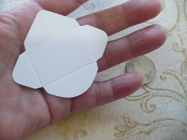 May include: A white folded card envelope, approximately the size of a US quarter, is held in a hand.