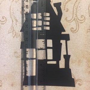 Op de afbeelding: Zwarte papieren uitsnede van een spookhuis silhouet, ongeveer 12,7 cm hoog, tegen een liniaal.