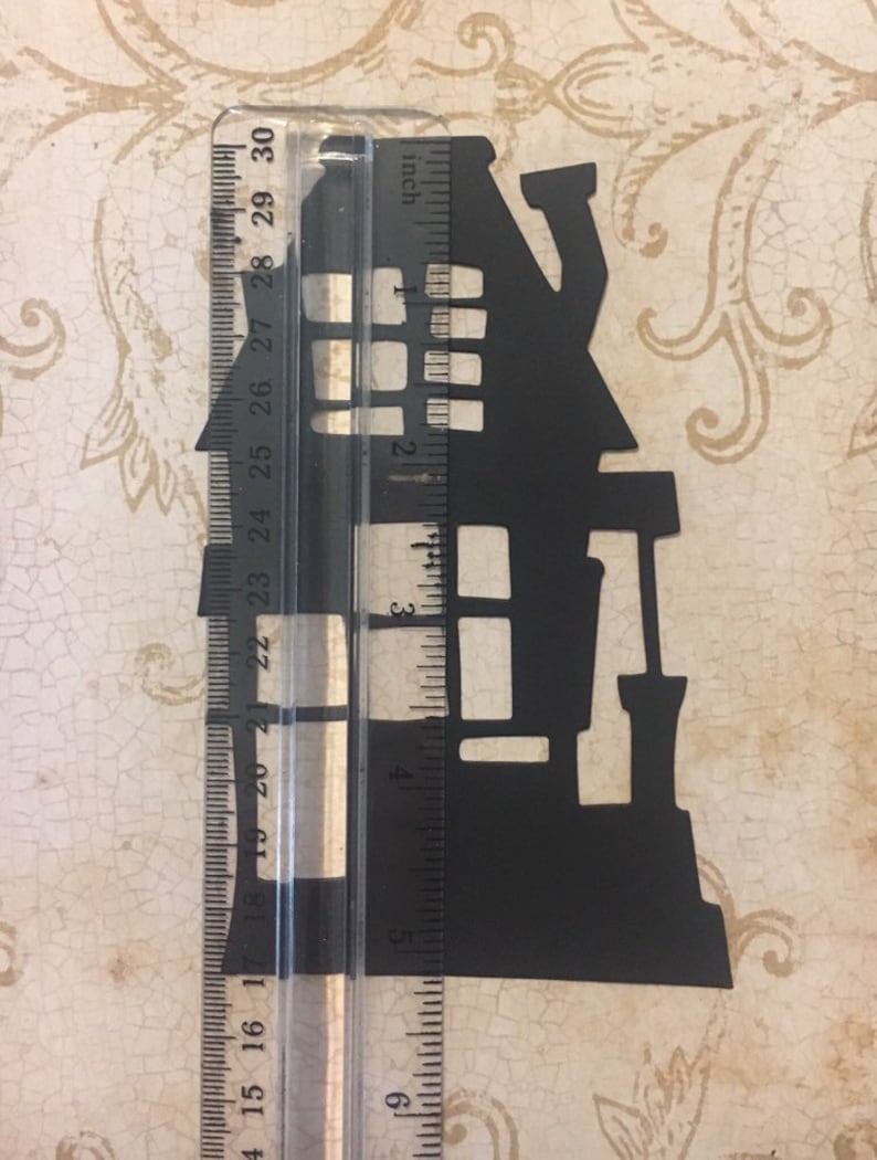 Op de afbeelding: Zwarte papieren uitsnede van een spookhuis silhouet, ongeveer 12,7 cm hoog, tegen een liniaal.