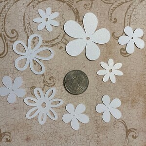 May include: Assortment of white paper flowers with a hole in the center. The flowers are various sizes and shapes, and are scattered on a textured brown surface.