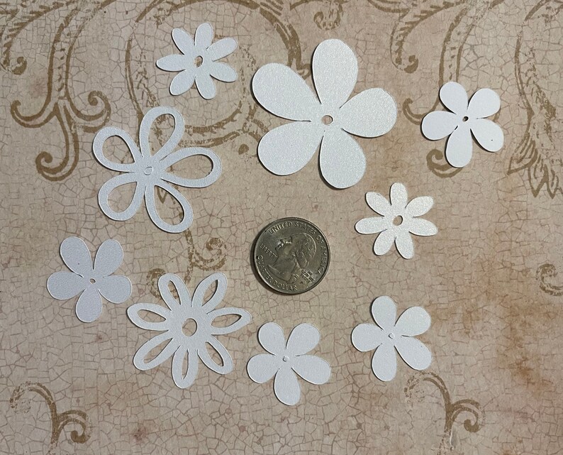 May include: Assortment of white paper flowers with a hole in the center. The flowers are various sizes and shapes, and are scattered on a textured brown surface.