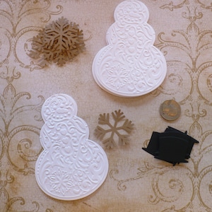 May include: Two white paper snowman ornaments with a decorative pattern. The ornaments are layered and have a 3D effect. There are two brown paper snowflakes and a black paper shape in the image.