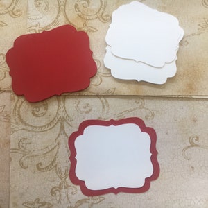 May include: Three red and white blank gift tags with a decorative scalloped edge. The tags are on a patterned background.
