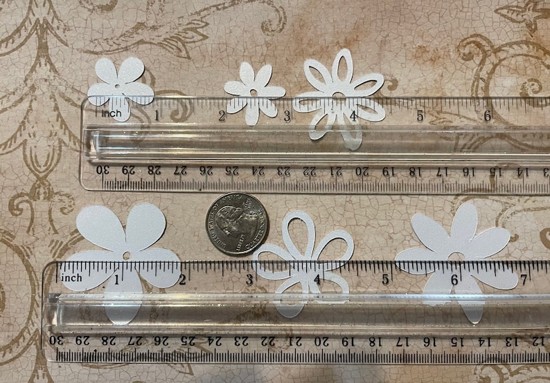 May include: White paper flower cutouts of various sizes, measured with a ruler in inches. The flowers are approximately 1 inch to 1.5 inches in diameter.