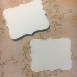 May include: Two white blank paper tags with a decorative scalloped border and a dotted line around the edge.