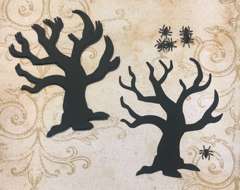 May include: Two black paper die-cut silhouettes of leafless trees with gnarled branches and five small black spider cutouts. The tree shapes feature thick trunks and spreading limbs, while the spiders are arranged in a small group and one alone.