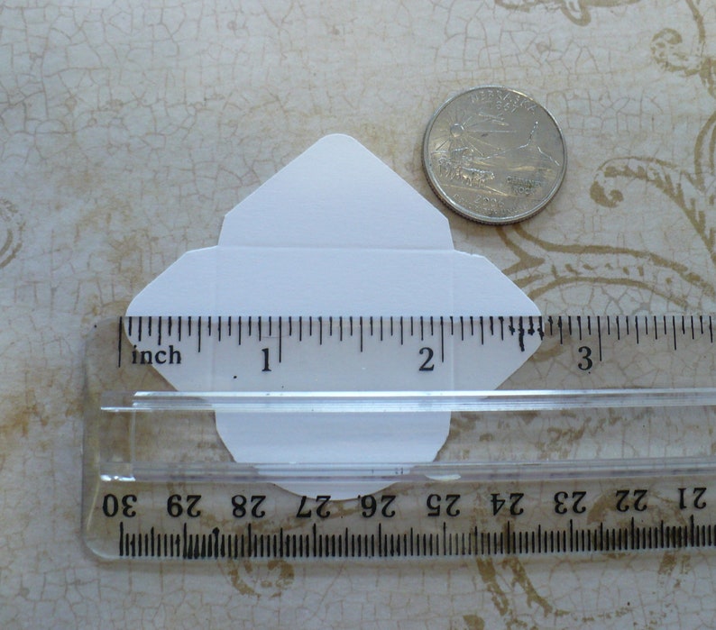 May include: A white envelope measuring 2 inches in length and 1.5 inches in width, next to a ruler and a US quarter.