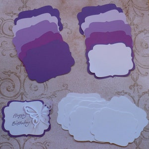 May include: A collection of purple and white decorative paper tags with scalloped edges. Some tags are solid purple in varying shades, while others are white. One tag features a cut-out butterfly and the handwritten text "Happy Birthday."
