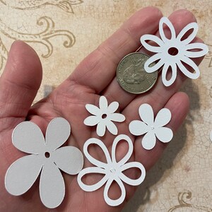 May include: A collection of five white paper flower cutouts of varying sizes. The flowers have a simple design with five petals and a small hole in the center.