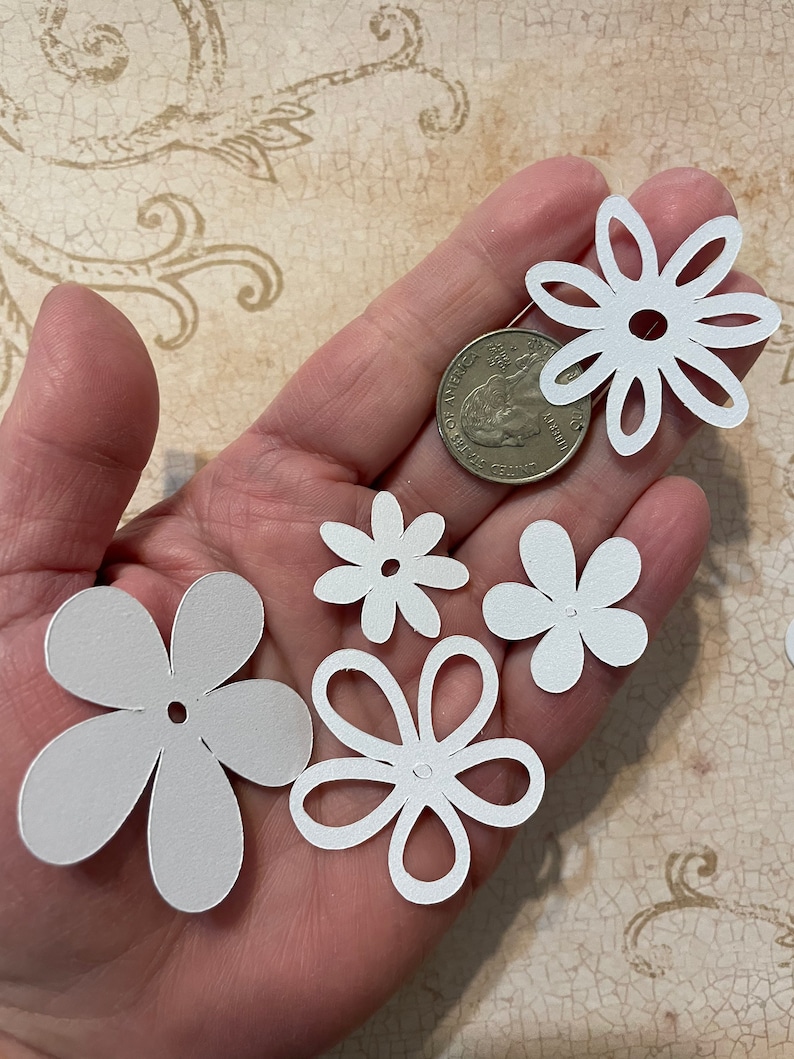 May include: A collection of five white paper flower cutouts of varying sizes. The flowers have a simple design with five petals and a small hole in the center.