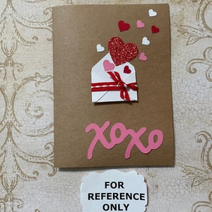 May include: A brown card with a white envelope with a red ribbon and a red glitter heart. There are pink and white hearts scattered around the envelope. The card says "xoxo" in pink letters.