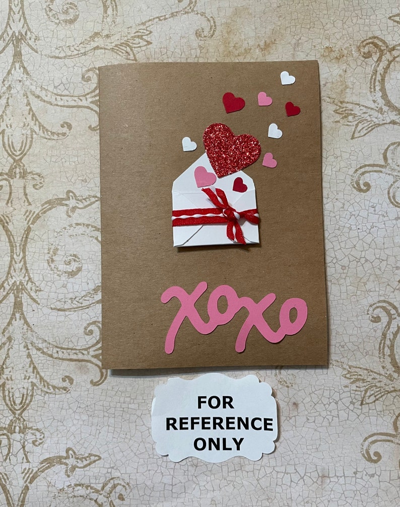 May include: A brown card with a white envelope with a red ribbon and a red glitter heart. There are pink and white hearts scattered around the envelope. The card says "xoxo" in pink letters.