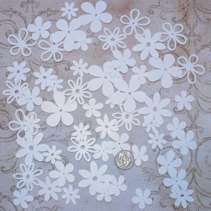 May include: A collection of white paper flowers of various sizes and shapes. The flowers are scattered on a textured, light brown surface.