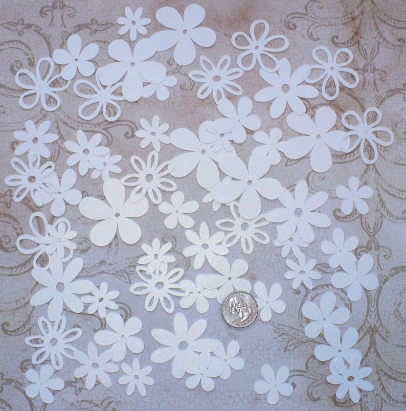 May include: A collection of white paper flowers of various sizes and shapes. The flowers are scattered on a textured, light brown surface.