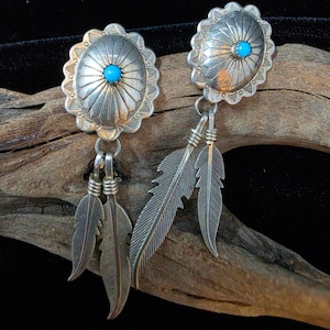 May include: A pair of silver earrings featuring round, scalloped-edge concho discs with a central turquoise stone. Each earring is adorned with two dangling silver feathers attached by small rings, showcasing detailed etched lines and textures.