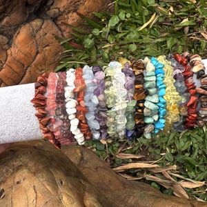 May include: A collection of colorful gemstone chip bracelets. The bracelets are made of various stones in shades of red, pink, white, purple, green, blue, and brown. The bracelets are displayed on a white cylindrical object, with a natural background.
