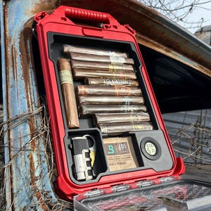 May include: A red, rectangular cigar humidor with a handle, open to reveal cigars, a lighter, and a humidity control pack. The humidor is set against a weathered, blue vehicle.