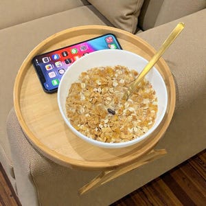 Könnte beinhalten: Ein runder Sofatablett aus Bambus hält eine weiße Schüssel mit Müsli, einen goldenen Löffel und ein Smartphone. Das Tablett ist für die Ablage auf einer Sofalehne konzipiert. Das Telefon zeigt App-Symbole an.