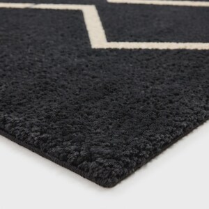May include: A dark charcoal gray rug with a thick, textured pile surface. The rug features a prominent, clean-lined geometric pattern in off-white, consisting of interconnected zigzag lines that form a repeating diamond-like shape across the fabric.