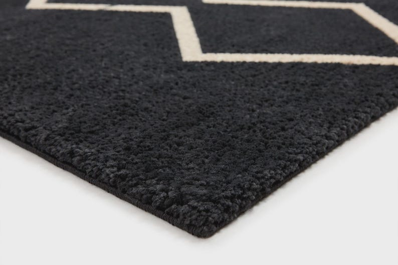 May include: A dark charcoal gray rug with a thick, textured pile surface. The rug features a prominent, clean-lined geometric pattern in off-white, consisting of interconnected zigzag lines that form a repeating diamond-like shape across the fabric.