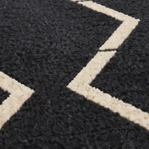 May include: A dark charcoal gray rug with a thick, high-pile, textured surface. The rug features a geometric pattern composed of bold, cream-colored lines that form angular shapes across the dark, fuzzy, and dense material of the textile.