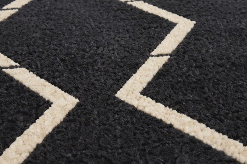 May include: A dark charcoal gray rug with a thick, high-pile, textured surface. The rug features a geometric pattern composed of bold, cream-colored lines that form angular shapes across the dark, fuzzy, and dense material of the textile.