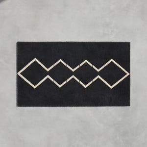 May include: A rectangular black doormat featuring a centered, continuous geometric pattern of four connected diamond shapes outlined in a light beige color. The mat has a textured, fibrous surface and a clean, straight-edged rectangular border.